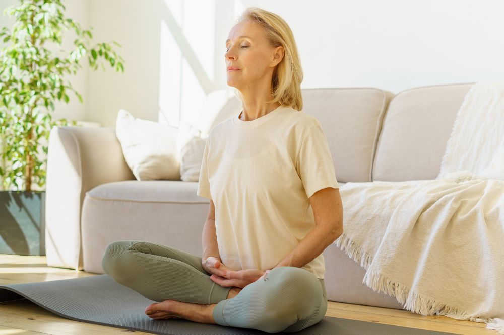 Žena v pohodlnom oblečení sedí na zemi v meditačnej pozícii, pretože meditácia je skvelý spôsob ako zatočiť so stresom.