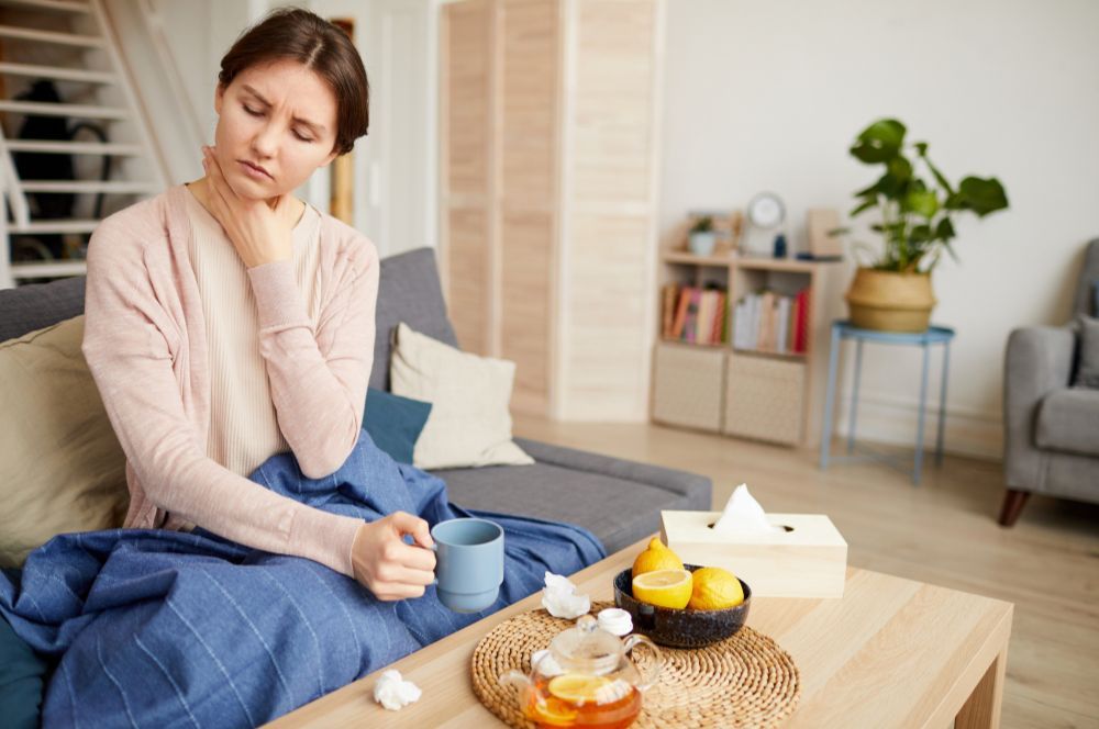 Žena sedí na gauči, pred sebou má čaj a servítky a bolí ju hrdlo, čo vyvolal vírus alebo baktéria.