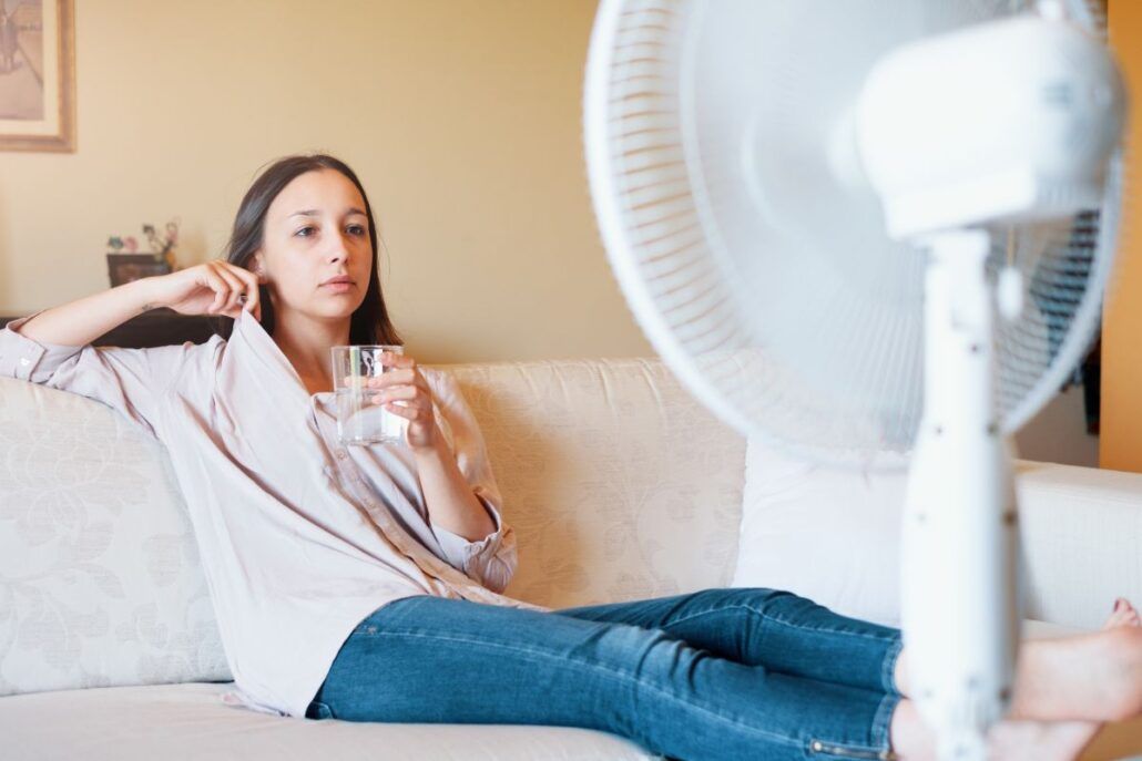 Žena sedí na gauči a ochladzuje sa ventilátorom.