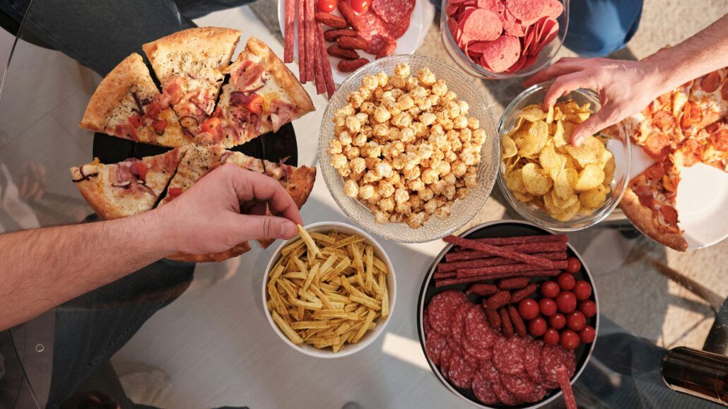 Na stole je položené rôzne nezdravé jedlo ako pizza, chipsy, salámy v miskách, ktoré vedú k nadváhe a tuku uloženému v oblasti brucha, ktoré sú rizikovým faktorom pre vznik inzulínovej rezistencie.