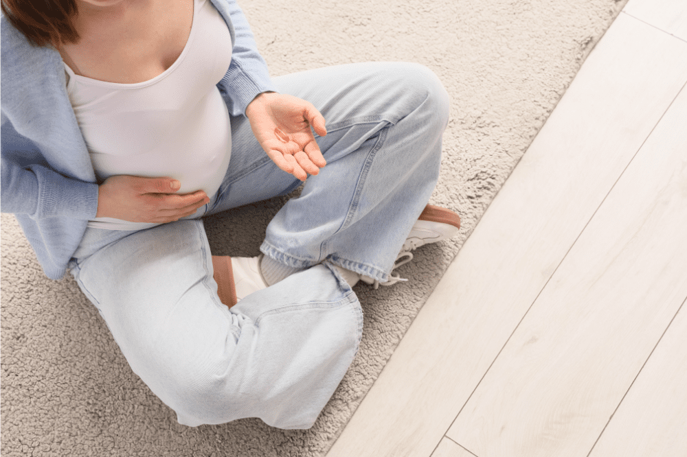 Tehotná žena sedí na zemi a v ruke ma vitamíny s kyselinou listovou, pretože dostatočný príjem kyseliny listovej sa odporúča ženám pred a počas tehotenstva, pretože deficit vitamínu B9 počas gravidity môže viesť k nenávratnému poškodeniu plodu