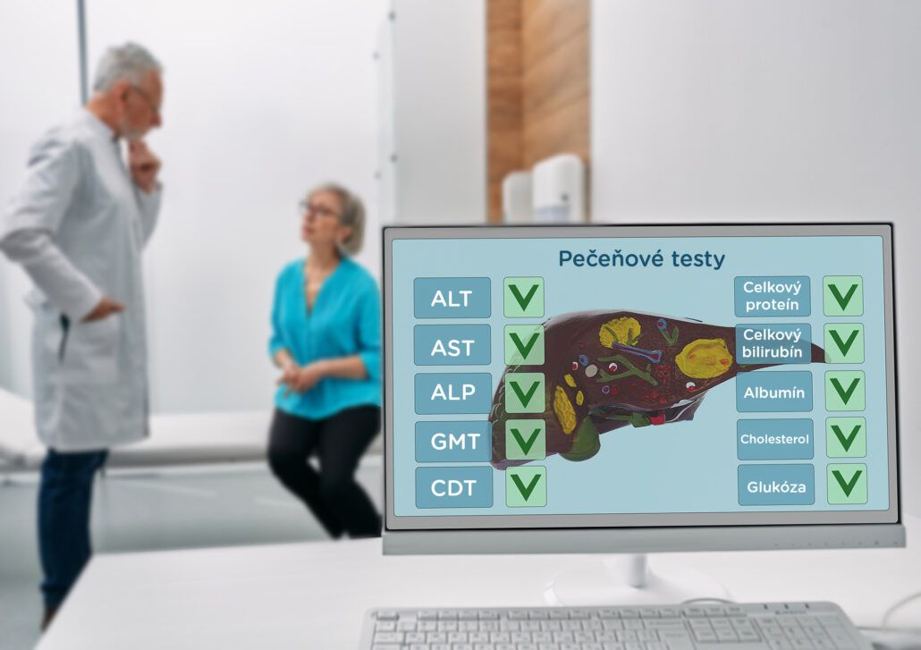 Na obrazovke počítača je vyhodnotenie pečeňových testov a lekár v pozadí vysvetľuje pacientke, ako dopadli.