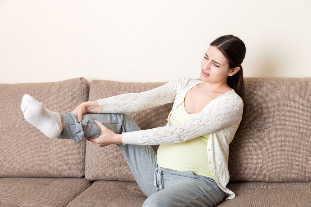 Tehotná žena sedí na pohovke a drží si nohu pretože cíti silné svalové kŕče.