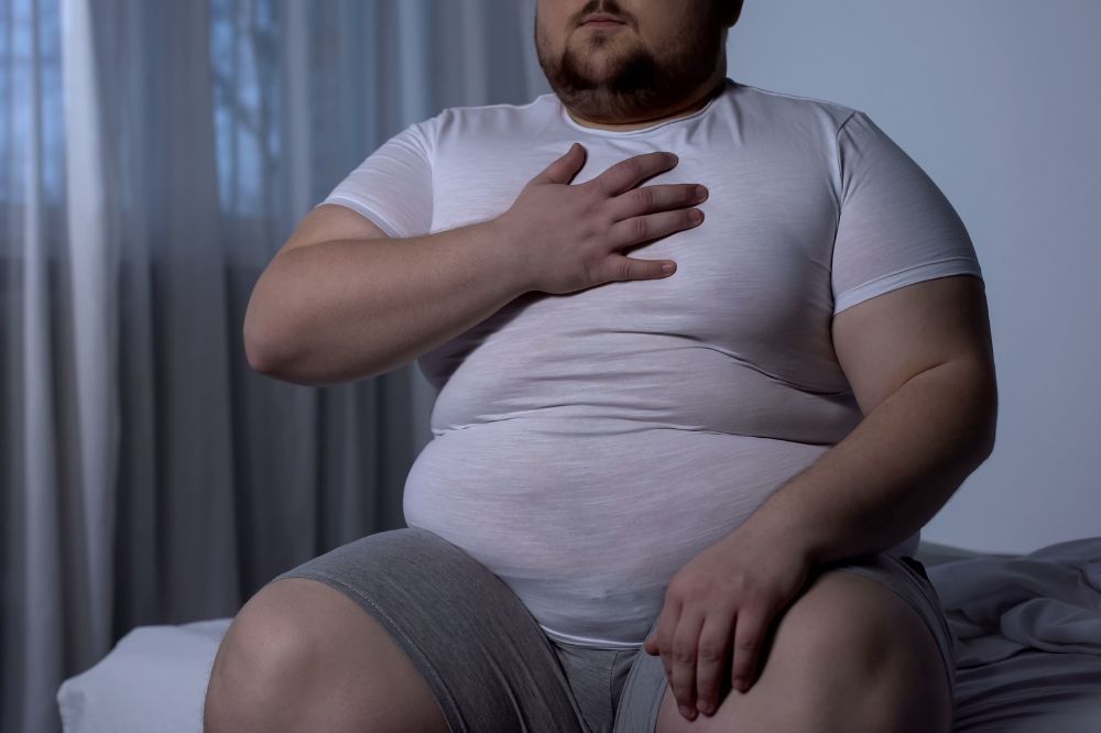 Obézny muž sedí na posteli a drží si ruku na srdci v dôsledku vysokého cholesterolu.