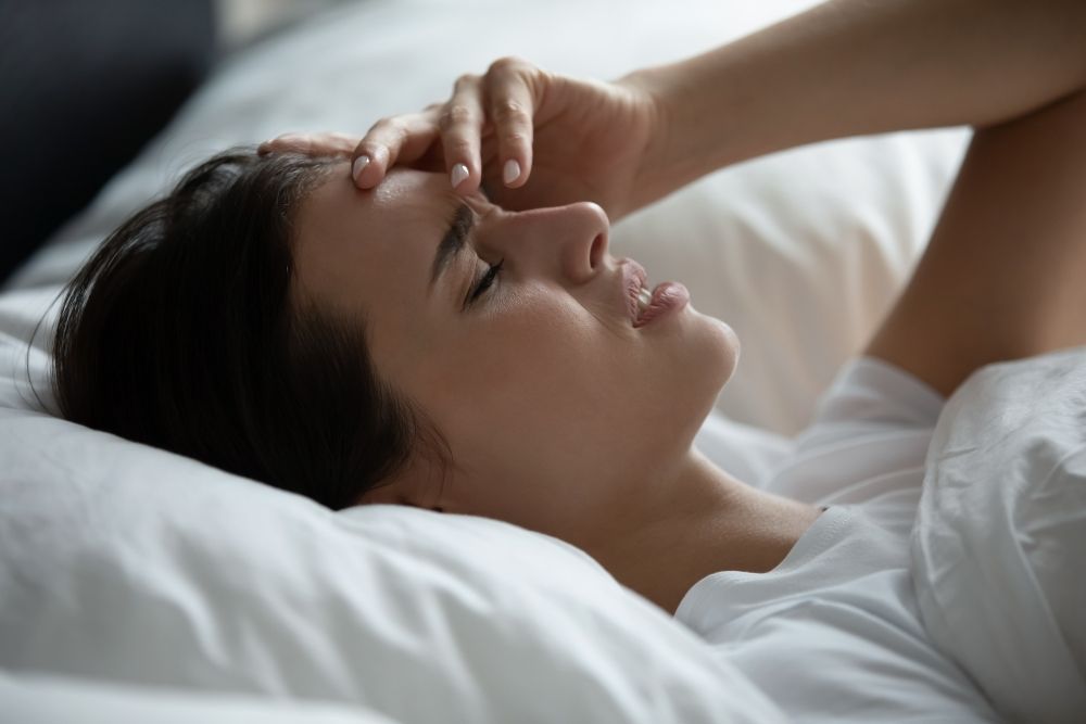 Pitný režim - dehydratácia, príznaky: žena s bolesťou hlavy leží v posteli a drží sa za čelo.