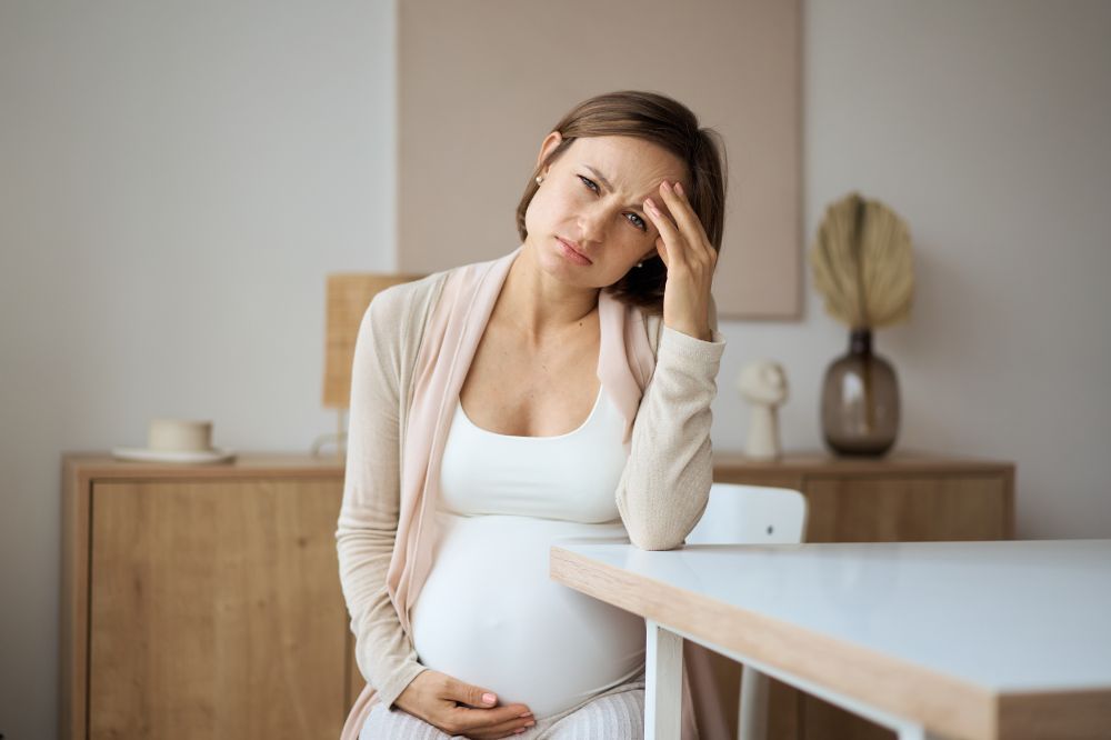 Tehotná žena sedí pri stole a drží si hlavu, pretože v rámci liečby preeklampsie je potrebné predčasný pôrod.
