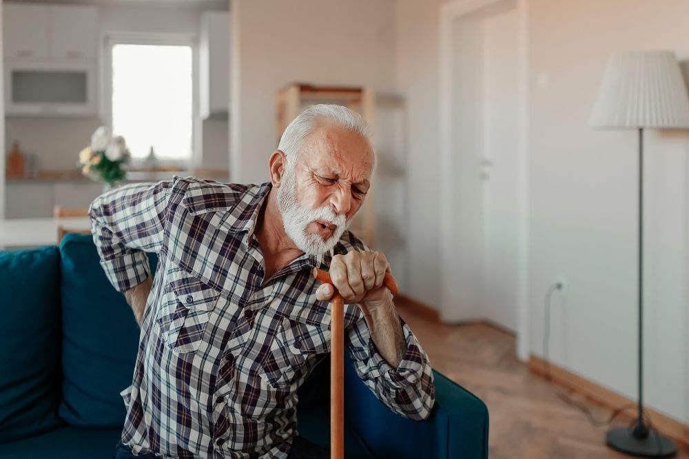 Bolesť chrbta: Starší pán s bradou opierajúci sa o paličku sa drží rukou v oblastí bedier.