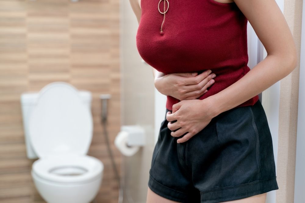 Problémy s trávením. Žena v červenom tričku sa drží za brucho a za ňou je wc