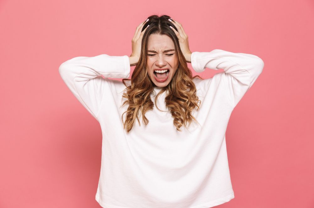 Žena v bielom tričku sa drží oboma rukami za hlavu, pretože dlhodobý stres a psychická nepohoda jej spôsobuje bolesť hlavy.