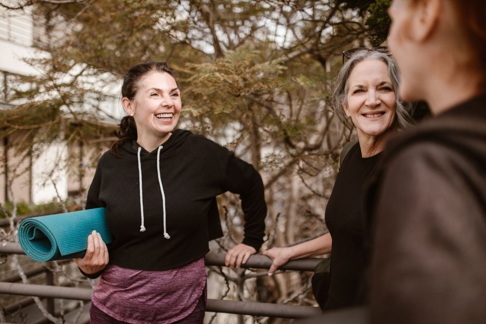 Prekonanie výkyvov nálad. Staršia žena drží v ruke podložku na cvičenie a usmieva sa na ďalšie dve ženy.