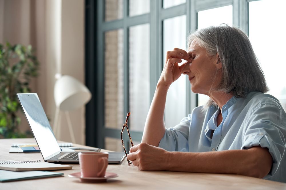 Staršia žena sedí pred počítačom a má zavreté oči v dôsledku únavy.