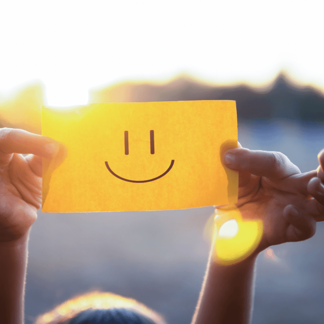 Dve ruky držia žltý papierik na ktorom sú nakreslené oči a úsmev ako znak šťastia.