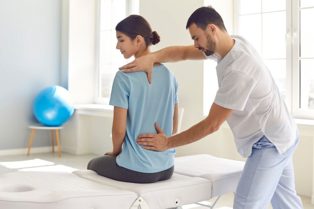 Vertebrogénny syndróm – vyšetrenie. Žene v modrom tričku sediacej na lôžku vyšetruje lekár chrbticu.