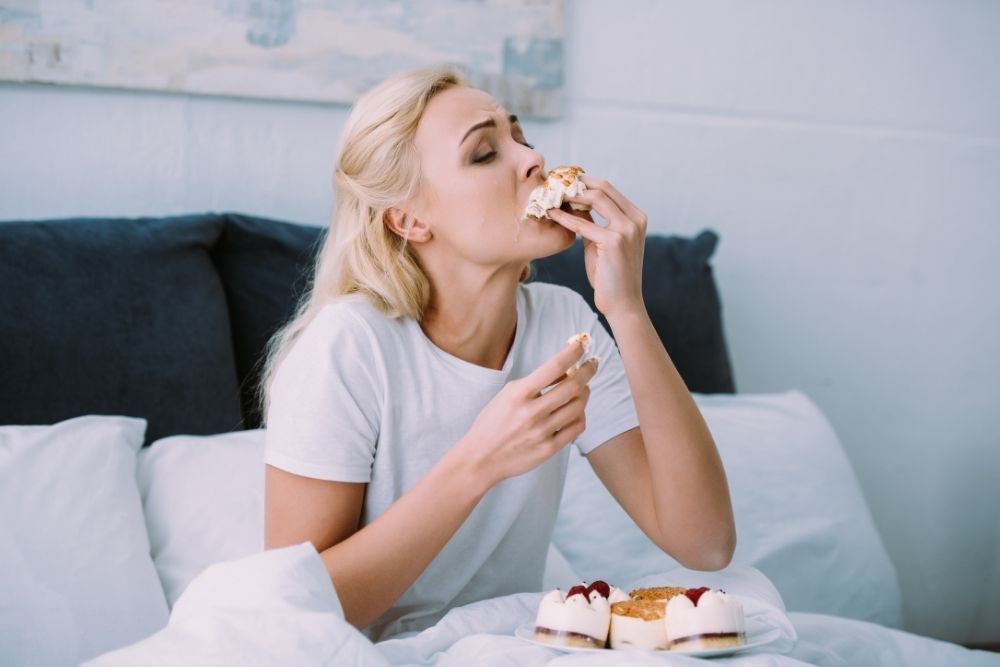 Pitný režim - dehydratácia príznaky: žena v bielom tričku sedí na posteli a s neovládateľnou chuťou je zákusky.