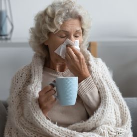 Staršia žena smrká do kapesníka a pije čaj, pretože má slabú obranyschopnosť a preto by mala brať GS Vitamín Lipo C 500 so šípkami.